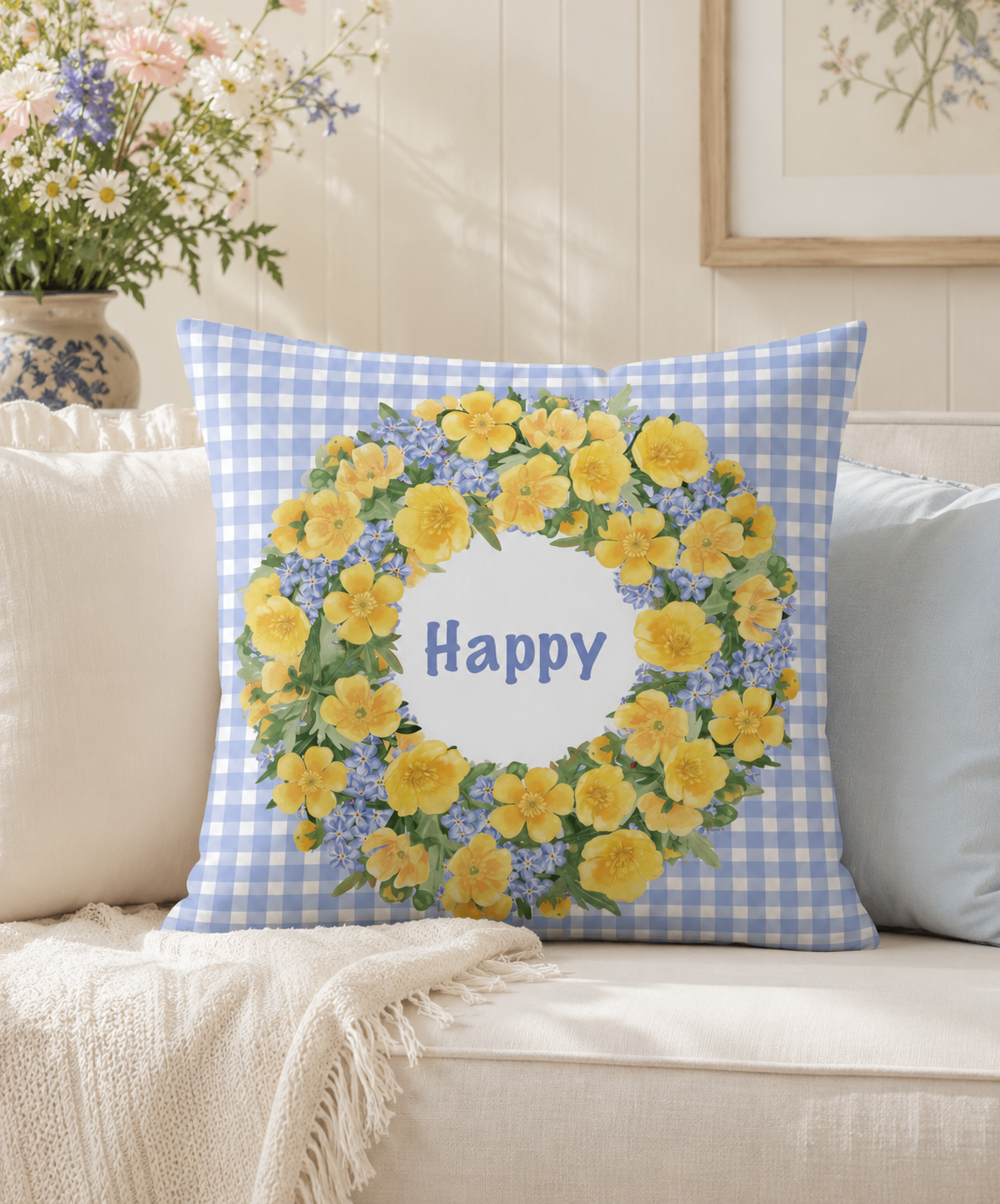 Blue gingham throw pillow with a buttercup wreath "Happy" design, resting on a cream sofa