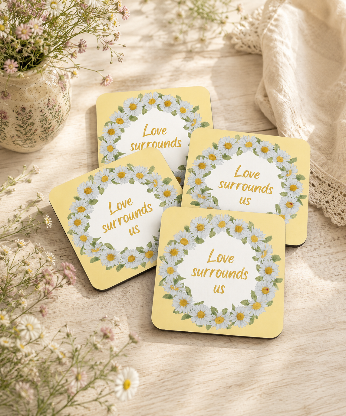 Set of four yellow daisy wreath coasters with "Love Surrounds Us" quote by Gabrielle Marlow, styled with wildflowers on a wooden table