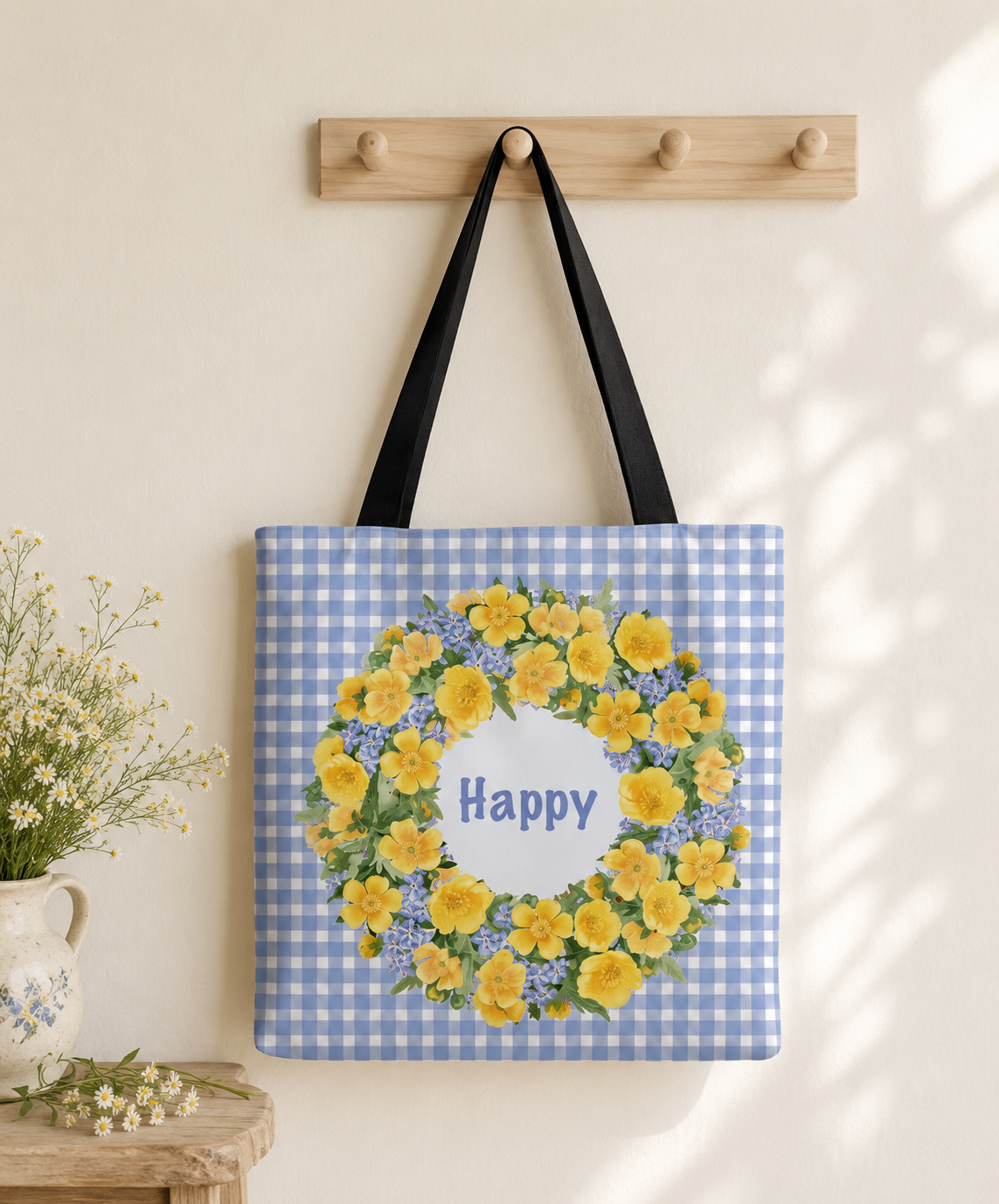 Blue gingham canvas tote bag with a yellow buttercup wreath and "Happy" design, hanging from a wooden hook