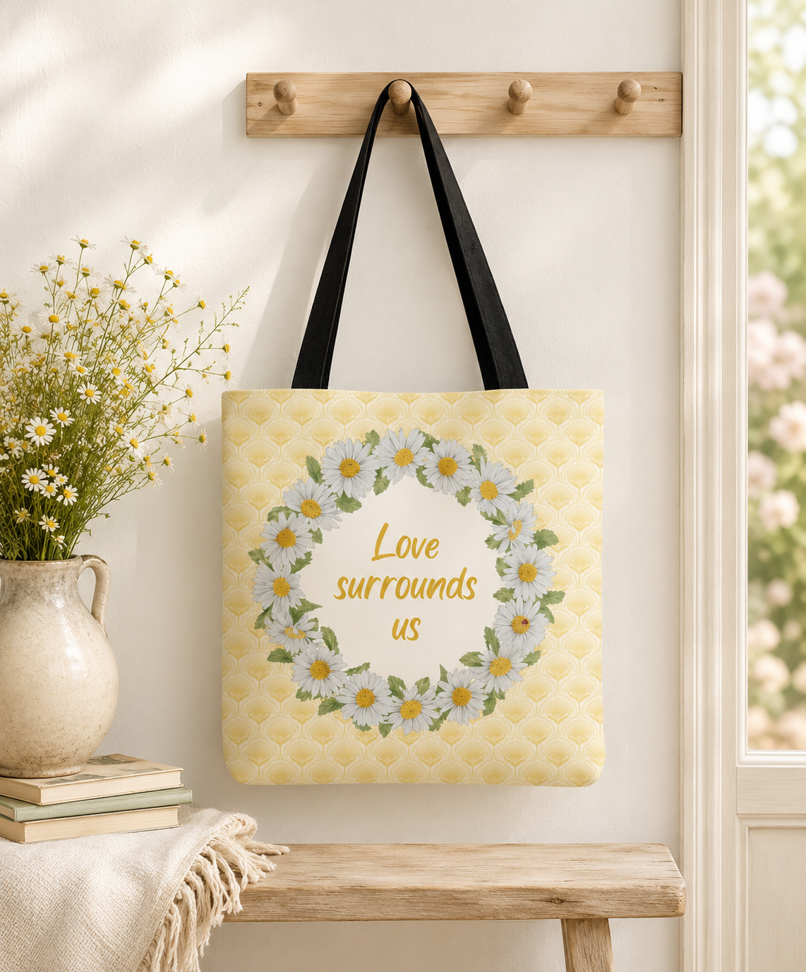 Yellow tote bag with white daisy wreath design and "Love surrounds us" quote hanging on a wooden hook, by Gabrielle Marlow